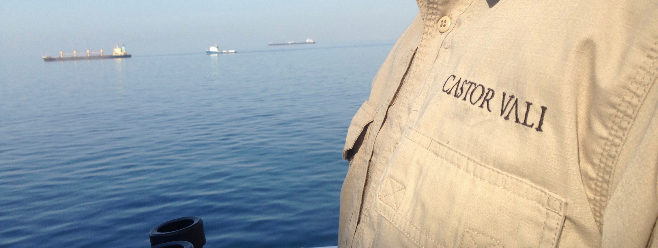Man on board a ship in Castor Vali branded shirt holding binoculars, infront of three ships at sea.